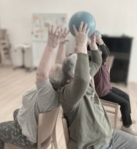 Abgebildet ist eine Partnerübung auf Stühlen, die mit der Rückenlehne aneinandergestellt sind. Ein Ball wird über Kopf übergeben.